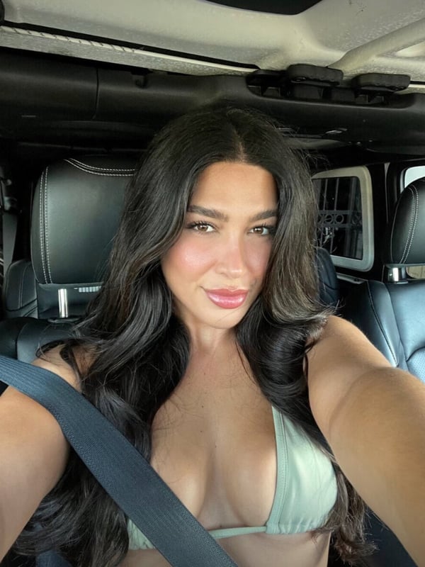 A woman sitting in a car interior, smiling at the camera while wearing a seatbelt and a green top.