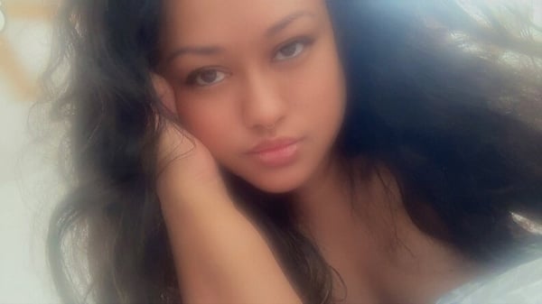 A young woman with long dark hair gazes thoughtfully at the camera in a softly lit, blurred portrait.