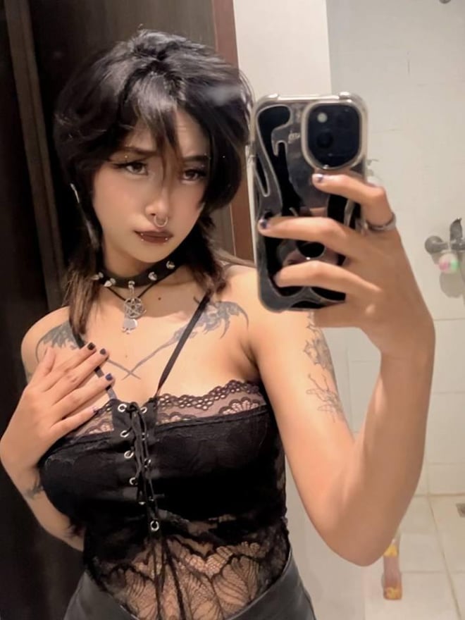 A woman with black hair, piercings, and tattoos poses topless in an indoor setting, cupping her chest.