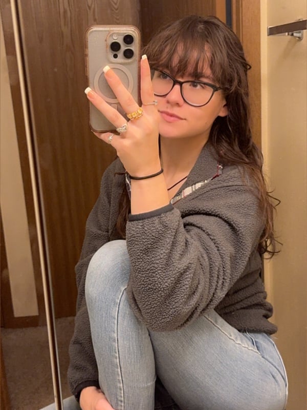 A young woman with glasses and long brunette hair takes a mirror selfie, sitting on the floor wearing a hoodie and jeans.