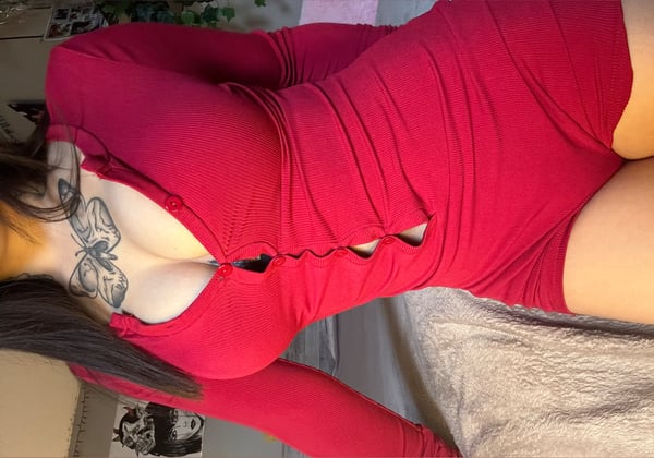 A woman posing on a bed in a red long-sleeve bodysuit, highlighting her figure against a cozy indoor background with plants.