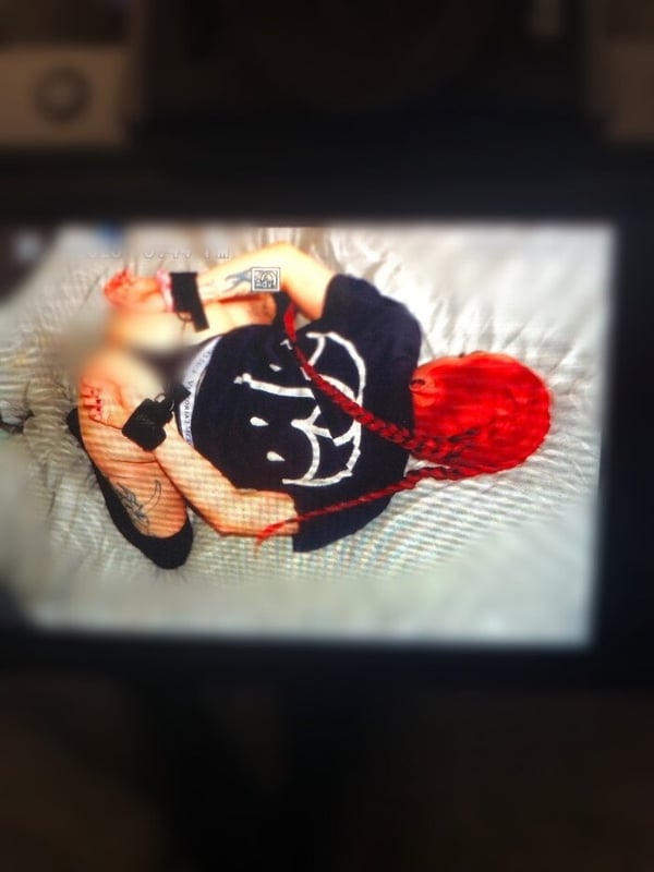 A woman with red hair lying on a bed, wearing black clothing and red ropes, with tattoos visible on her arms.