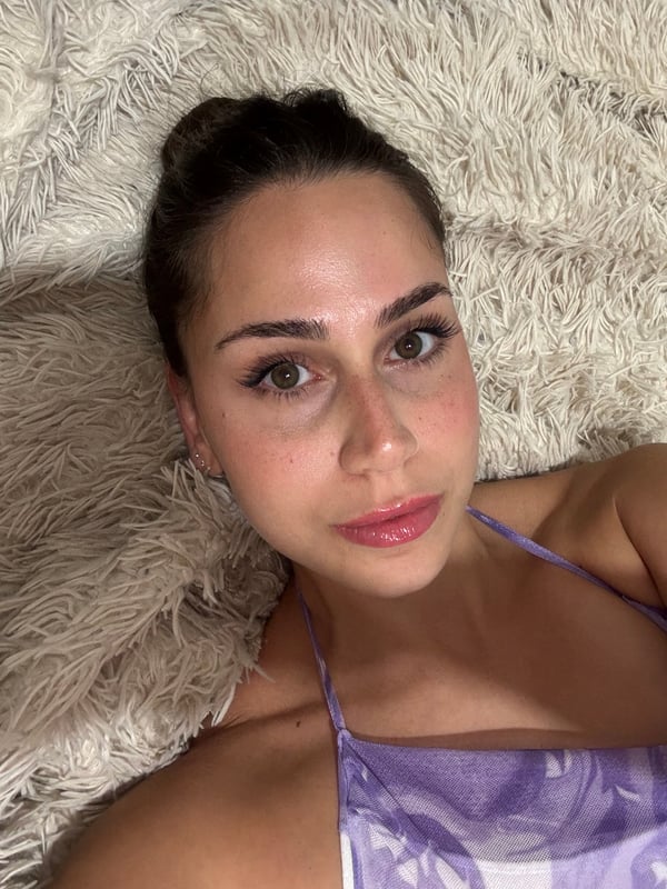 A close-up selfie of a young woman with dark hair pulled back, lying on a fluffy white surface and wearing a purple top.