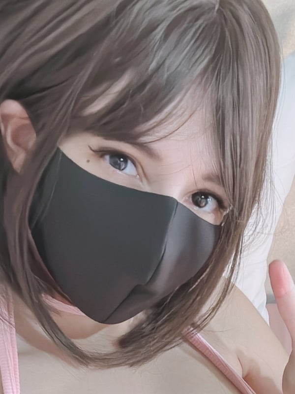 Close-up of a young woman with long brown hair wearing a black face mask, making a peace sign against a soft pink background.