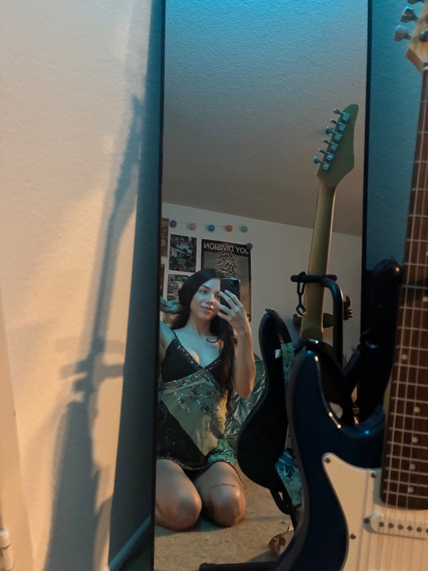 Young woman with long wavy dark hair smiling at the camera, wearing a gray long-sleeve crop top, standing in a colorful bedroom with posters on the walls.