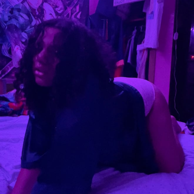 A young woman with curly hair posing on a bed in a colorful bedroom with artistic wall decor and neon lighting.