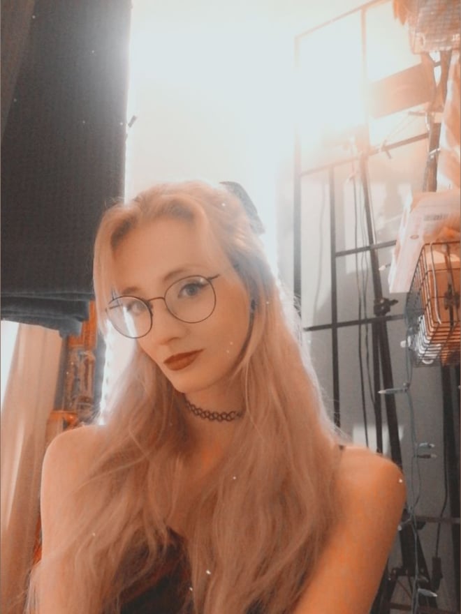Young woman with long blonde hair and glasses posing indoors with warm lighting and industrial background elements.