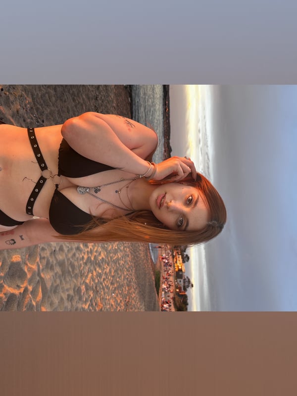 Young woman with red hair posing on a beach at sunset, wearing a bikini and jewelry.