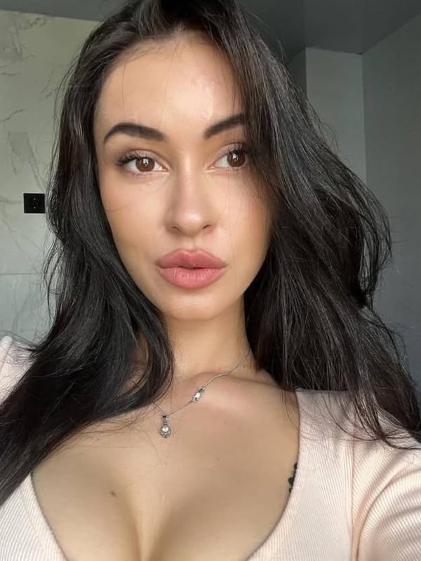 A woman with long dark hair takes a close-up selfie in a light-colored top, smiling softly in an indoor setting with frames on the wall.