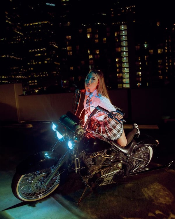 A woman posing confidently on a motorcycle at night with city lights and skyscrapers in the background.