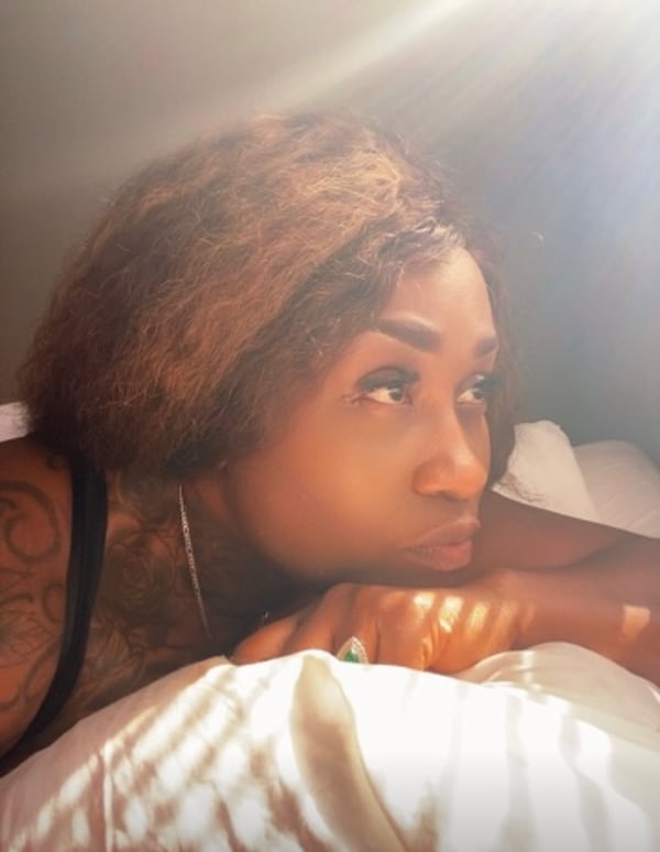 A woman with curly brown hair and tattoos poses thoughtfully on a bed with white sheets, illuminated by soft sunlight.