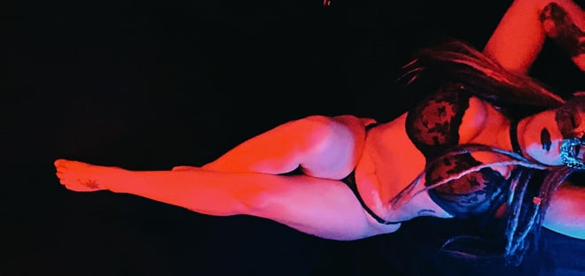 A woman reclining on the floor in black lace lingerie and a mask, illuminated by red and blue lighting against a dark background.
