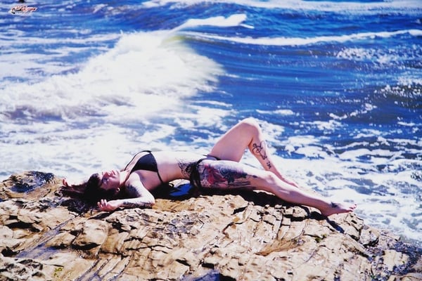 A woman in a black bikini lounging on rocks by the ocean with waves crashing nearby, showcasing tattoos on her legs.