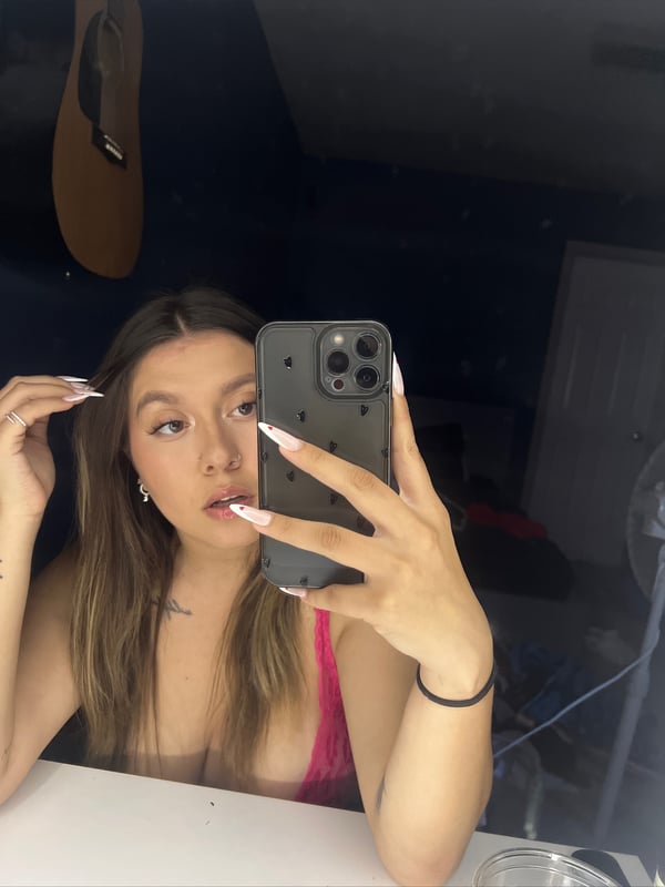 Young woman taking a selfie in a room, holding her phone with long nails, wearing a pink top.