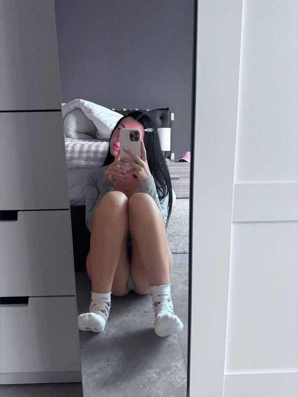 A young woman sitting on the floor in a bedroom, taking a mirror selfie while wearing socks and casual clothing.