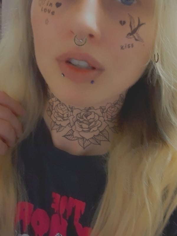 Close-up of a young woman with long blonde hair, blue eyes, facial piercings, and neck tattoos, posing indoors.