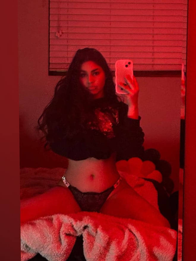 Young woman taking a selfie in a dimly lit room with red lighting, wearing a cropped top and sitting on a bed.