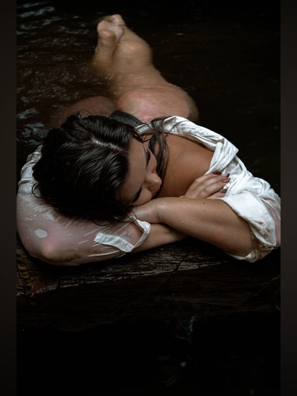 A close-up of a woman lying in shallow water on wood, wearing a wet white shirt, with her legs partially visible.