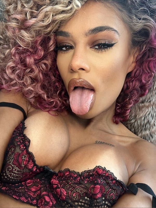 A woman with curly pink and blonde hair sticks out her tongue while posing in lace attire on a textured surface.