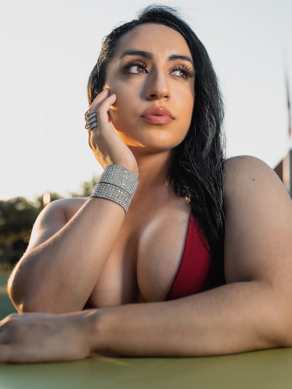 A young woman with long dark hair and striking eyes posing thoughtfully outdoors in a red top, accessorized with rings and bracelets.