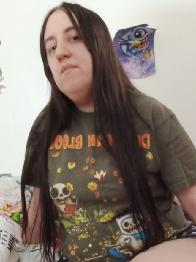 Young woman with long brown hair wearing a graphic t-shirt, sitting casually in a room decorated with posters and plants.