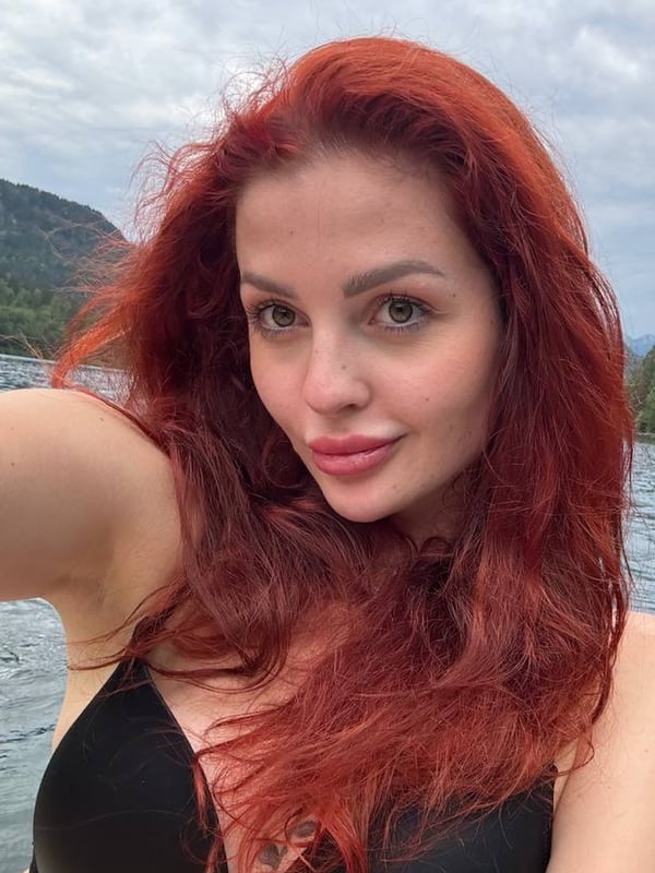 Young woman with long red hair taking a selfie by a scenic lake with mountains, wearing a black swimsuit top.