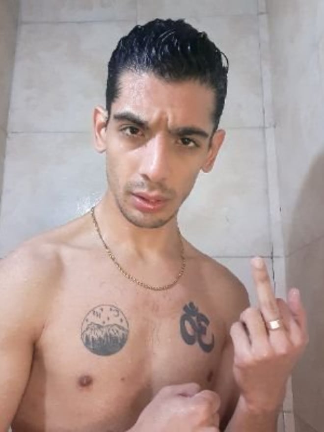 Shirtless man with tattoos standing in a tiled shower area, making a hand gesture while wearing a necklace.