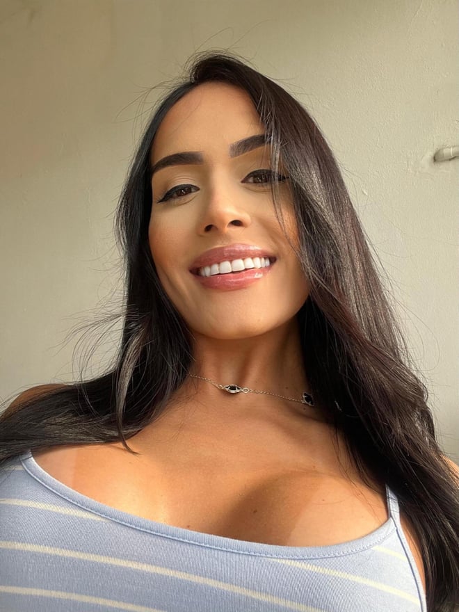 A woman with long dark hair smiling at the camera, wearing a light blue striped top, against a simple indoor background.