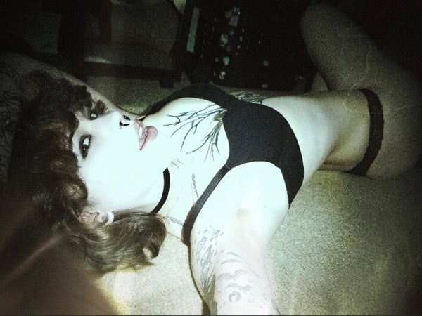 A young woman with curly hair reclines on a bed in dark lingerie, posing seductively with tattoos visible on her skin.