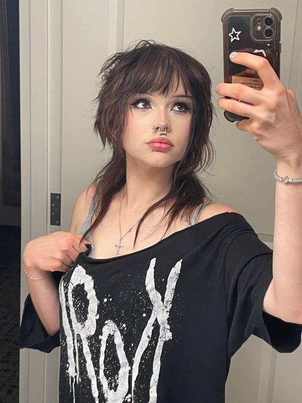 Close-up portrait of a person with dark hair and dramatic eye makeup posing with their hand near their face.