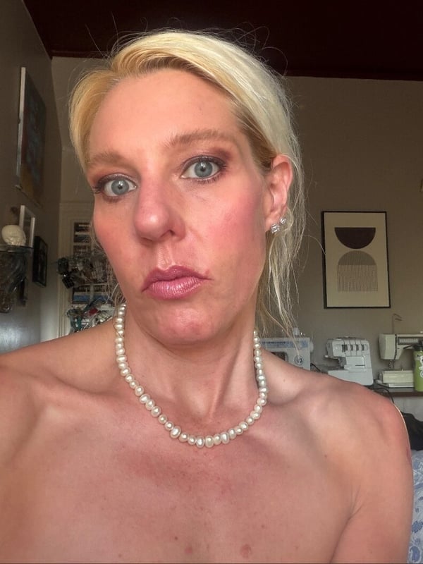 A close-up portrait of a blonde woman with blue eyes and pearl necklace, pouting slightly against a home interior background.