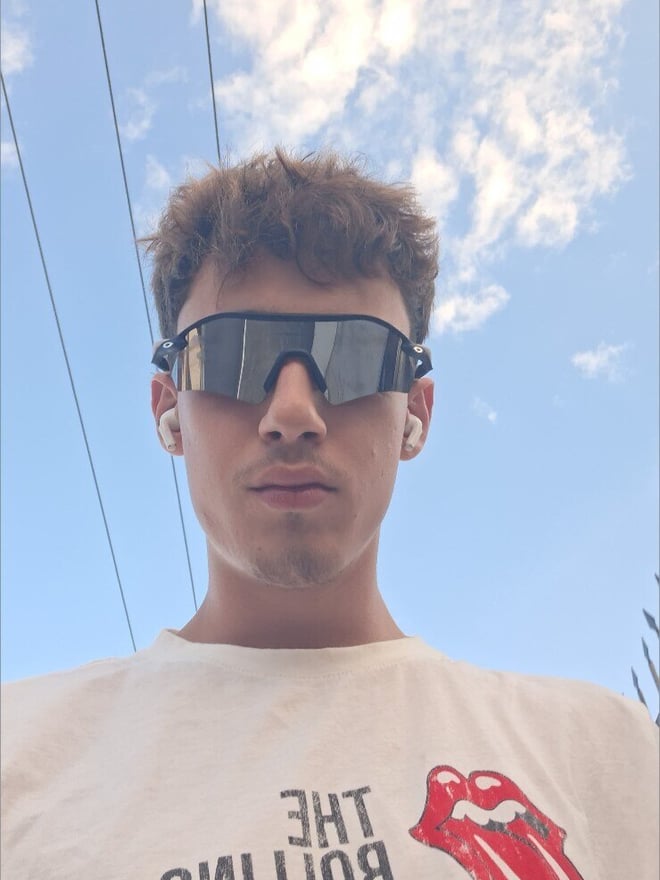 A young man wearing sunglasses and a Rolling Stones t-shirt, standing outdoors under a partly cloudy sky with power lines in the background.