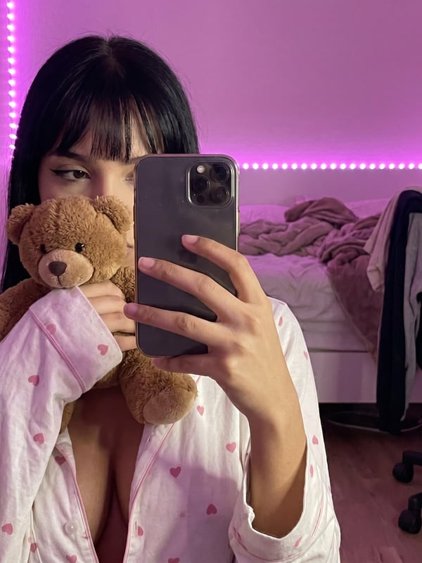 Young woman with long dark hair sitting on a bed in a pink bedroom, wearing a white tank top and pink shorts, holding her phone.
