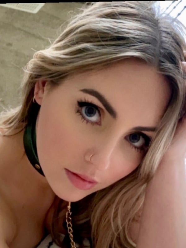 Close-up portrait of a woman with blonde hair, blue eyes, and a choker necklace, resting her head on her hand while lying down.