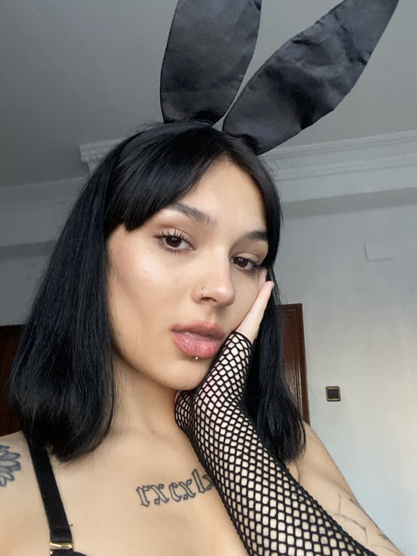A woman with dark hair wearing bunny ears and fishnet gloves, posing with a hand on her face indoors.