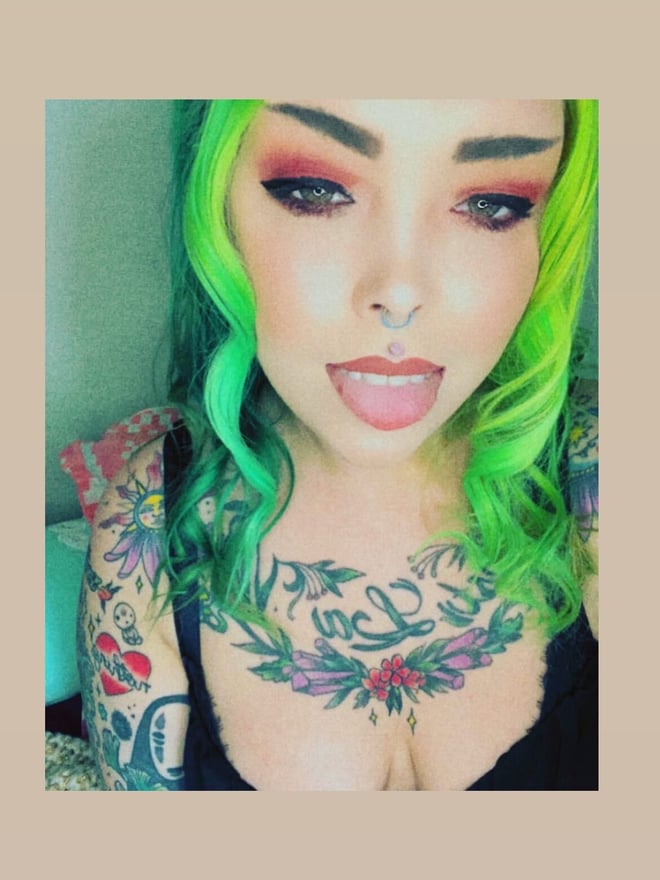 Close-up portrait of a woman with vibrant green hair, facial piercing, and extensive tattoos on her chest and arms, wearing a black top.