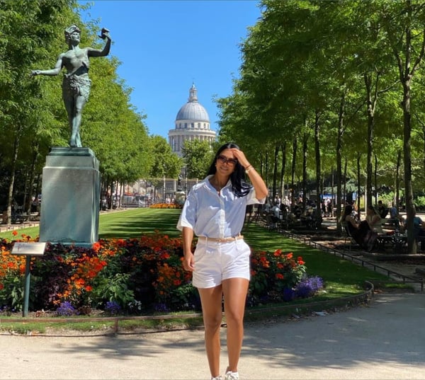 A woman in a white shirt and shorts posing in front of a statue and domed building in a lush park setting.