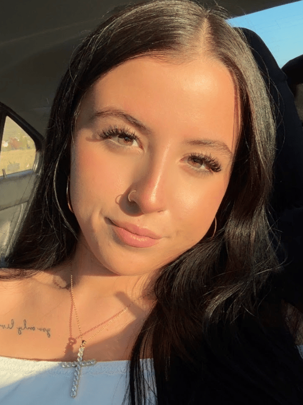 Young woman with long dark hair, wearing a white top and necklace, smiling in a car with sunlight streaming in.