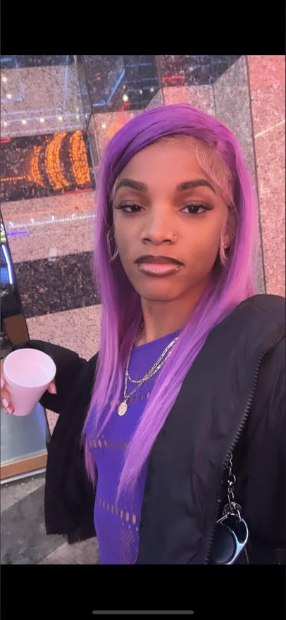 Young woman with long purple hair standing outdoors at night, holding a cup and wearing a dark jacket over a purple top.