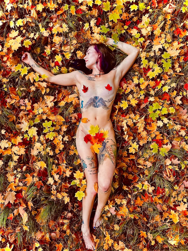 A woman with tattoos and purple hair lies topless on a bed of colorful autumn leaves outdoors.