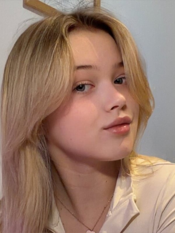 Close-up portrait of a young blonde woman smiling gently while wearing a light-colored top in a softly lit room.