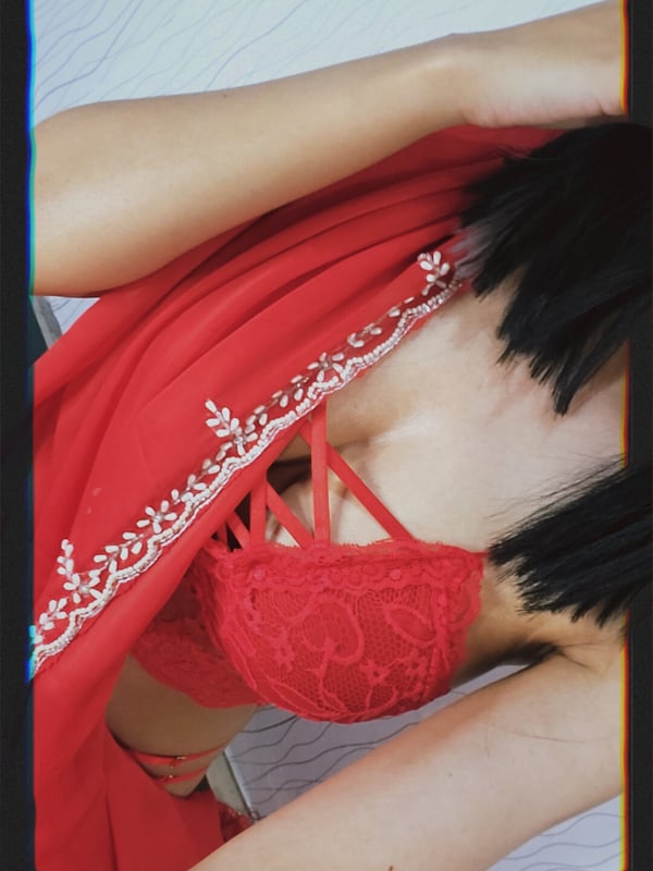 Woman in a red lace top and embroidered red garment posing on a bed with a blurred background.