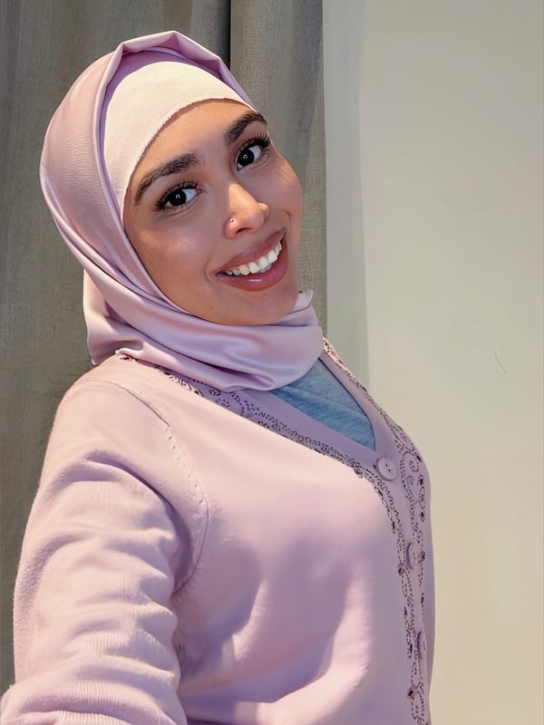 A smiling woman wearing a light purple headscarf and cardigan, taking a selfie with a slight tilt.