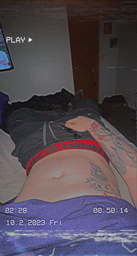 A young man lying on a bed in dark underwear, displaying tattoos on his abdomen and arm, in a dimly lit room with a TV in the background.