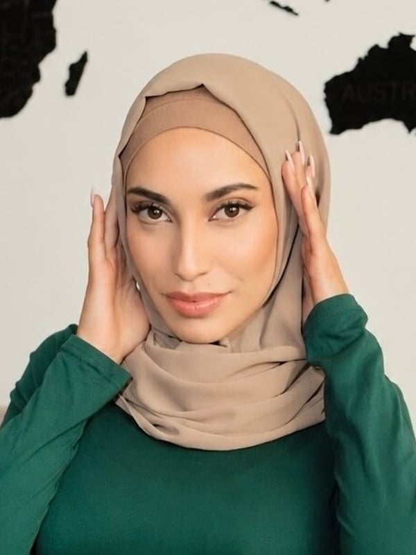 Woman in a beige hijab smiling and touching her face with both hands, posed in front of a large black map of Australia.