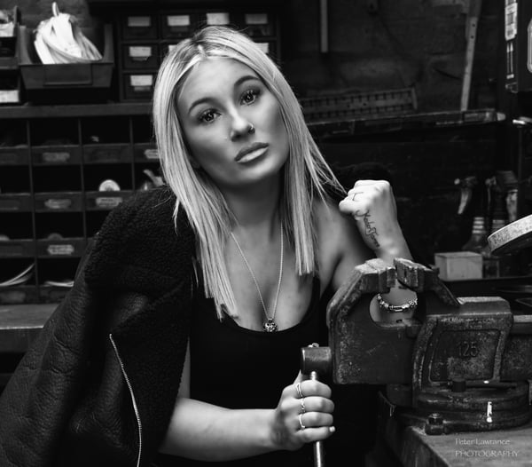A young woman with blonde hair posing confidently in a workshop setting surrounded by tools and shelves, wearing a dark top and jacket.