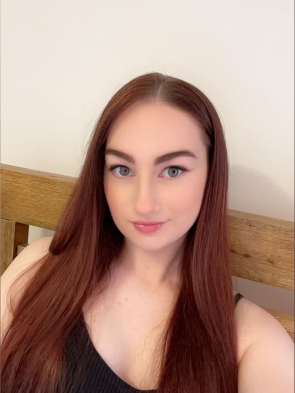 Young woman with long red hair smiling at the camera, wearing a black top, sitting in a bedroom with a wooden headboard.