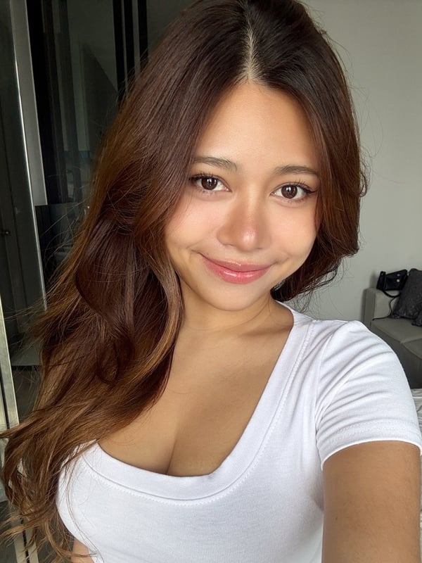 A smiling woman with long brown hair wearing a black top and necklace, seated on a bed.