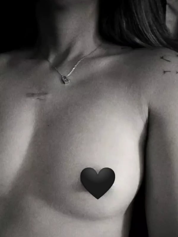 Close-up portrait of a woman with long hair and a necklace, featuring a black heart sticker on her upper chest against a dark background.