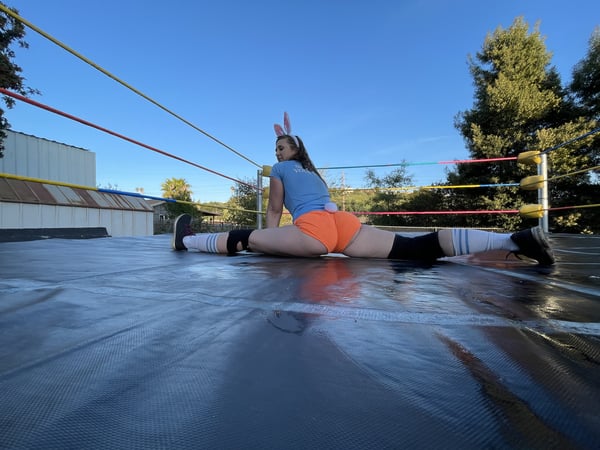 A woman in an orange outfit and bunny ears performs a flexible split pose inside a wrestling ring on a rooftop with trees and blue sky in the background.
